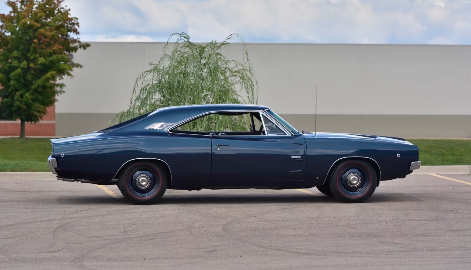 A classic dark blue muscle car is parked in an empty lot, viewed from the side. The background features a trimmed tree, a brick building, and a light-colored wall under a partly cloudy sky.