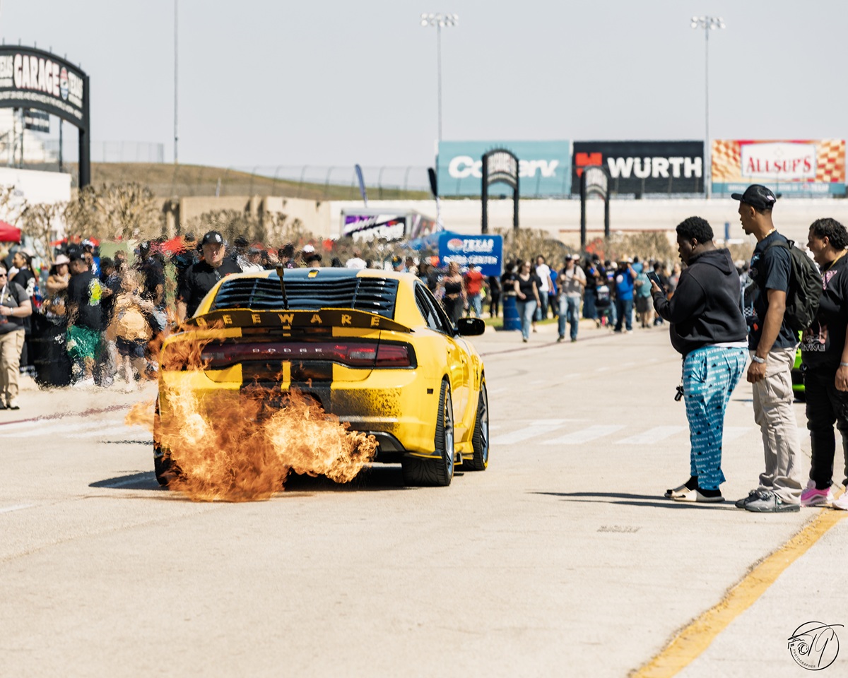 A yellow sports car emits flames from its exhaust as it drives on a racetrack, with onlookers standing nearby and crowds gathered along the track.