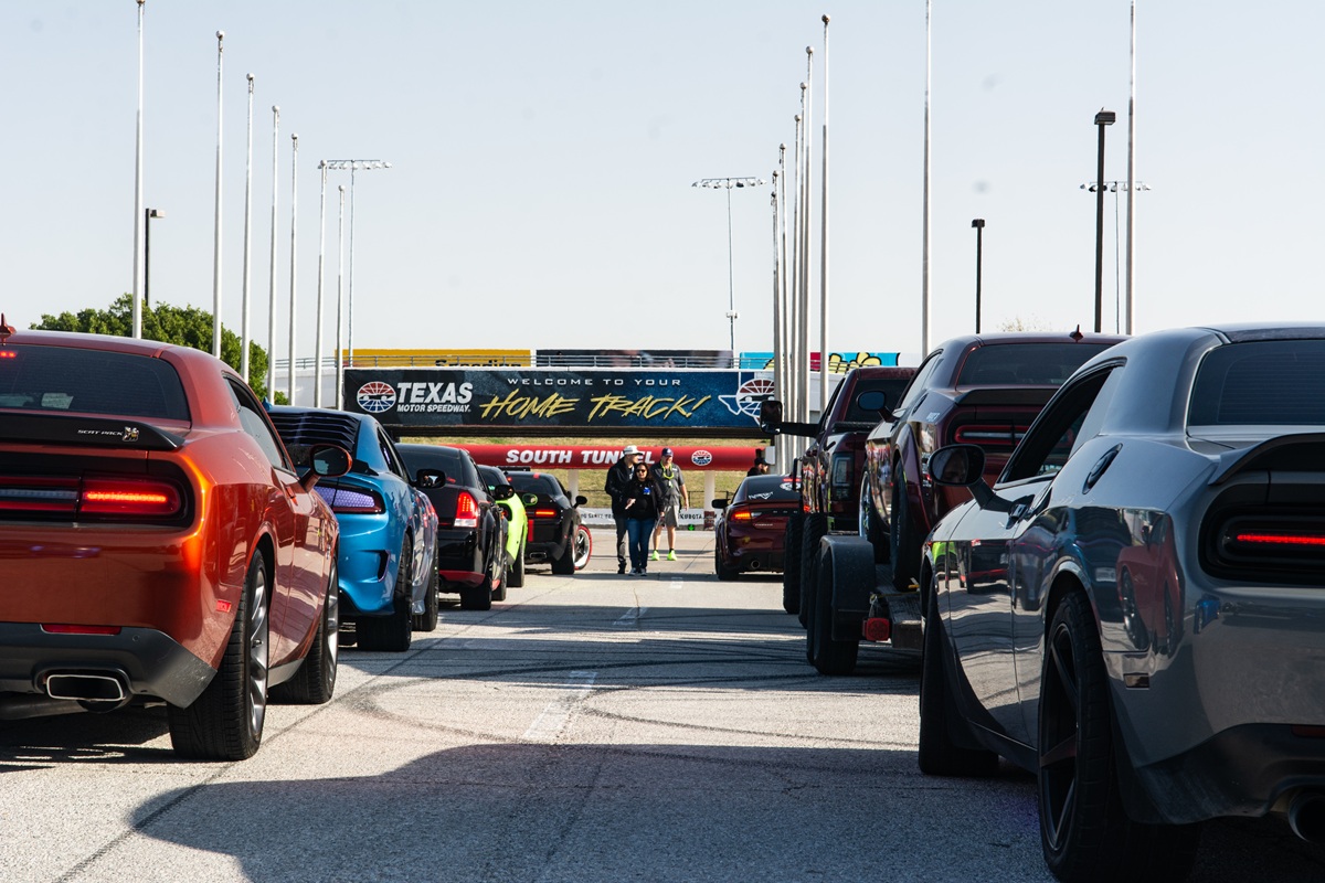 Several sports cars are lined up on a racetrack under clear skies, facing a sign that reads Welcome to your home track at Texas Motor Speedway, with a few people standing in the distance.