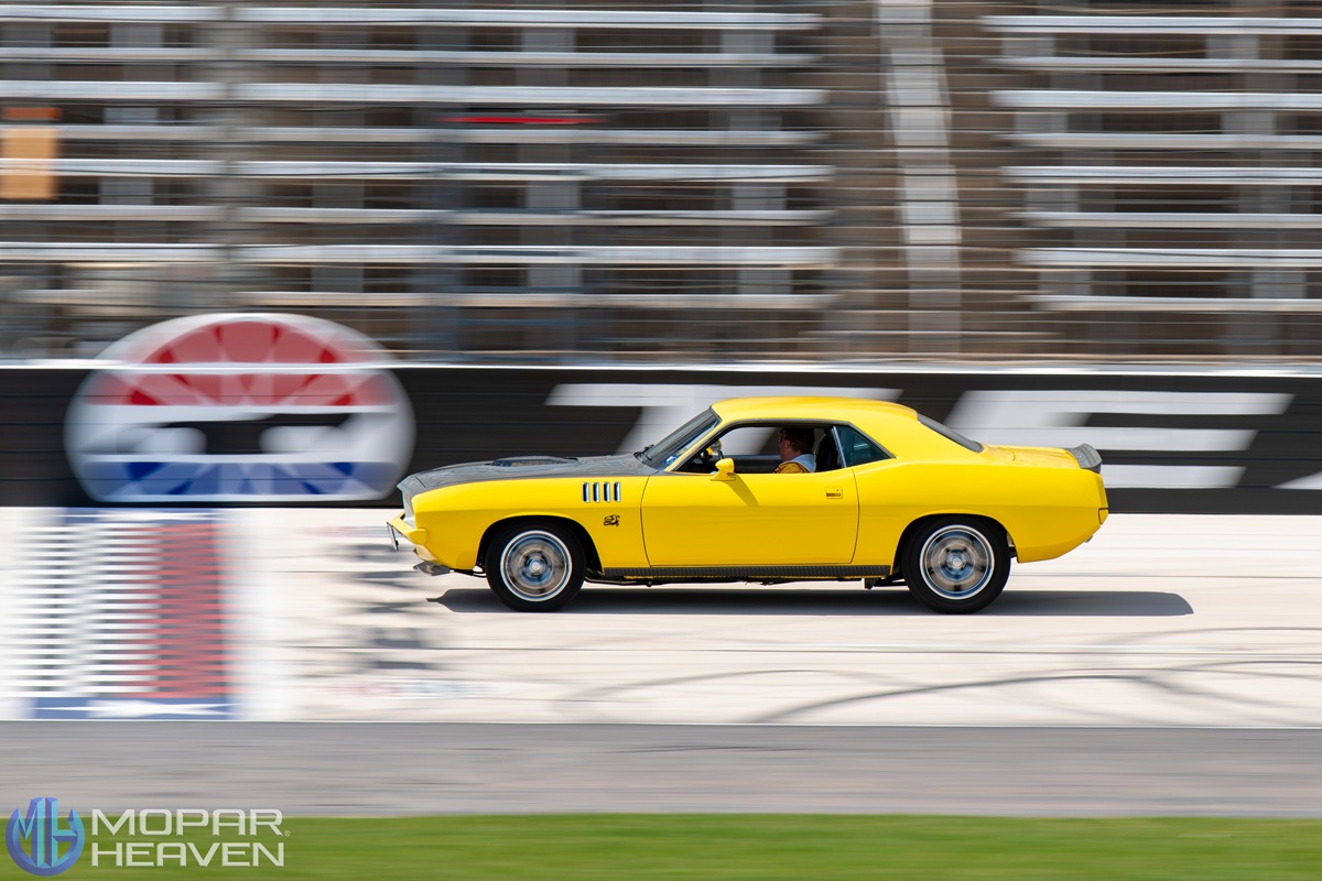A bright yellow classic muscle car speeds past empty bleachers on a racetrack, with motion blur in the background. The car is in sharp focus, and a logo for Mopar Heaven appears in the lower left corner.