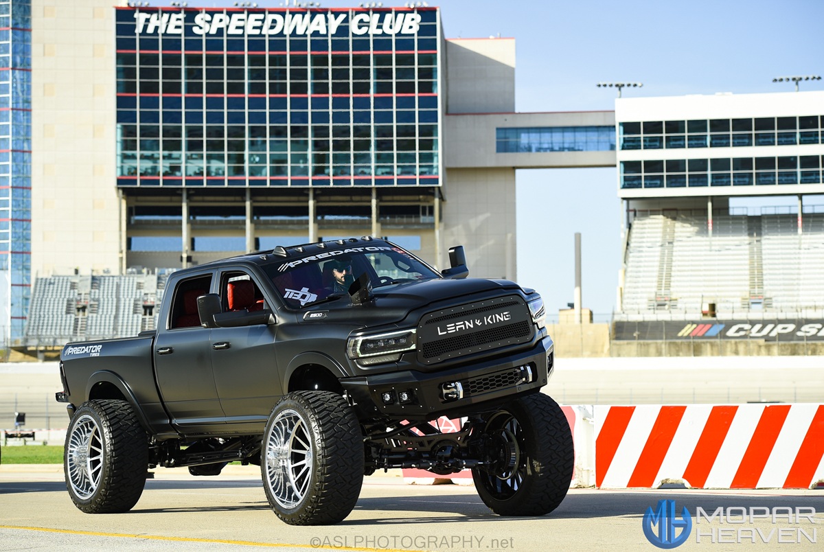 A lifted black pickup truck with large custom wheels is parked at a racetrack, with LEASH KING on the grille. The Speedway Club building and stadium seating are visible in the background.