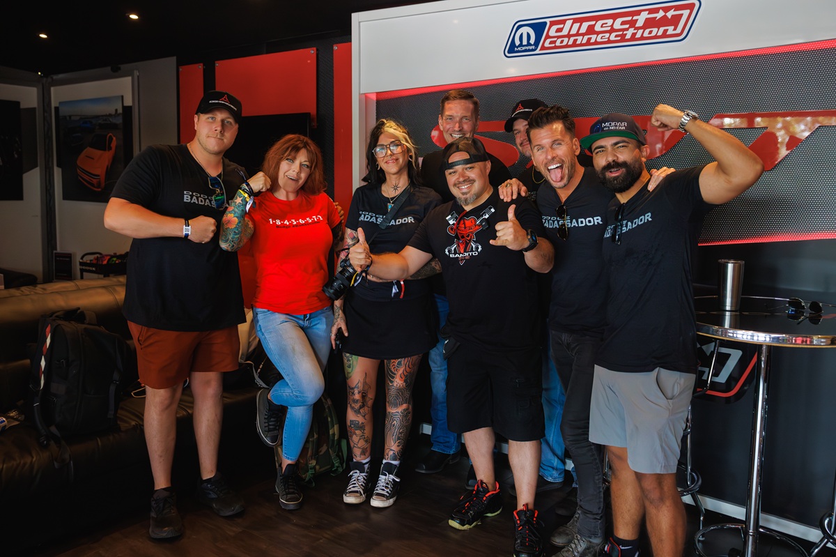 A group of seven people stand together indoors, smiling and posing enthusiastically. They wear casual clothing, some in black Badaad shirts, with automotive-themed decor and a Direct Connection sign in the background.