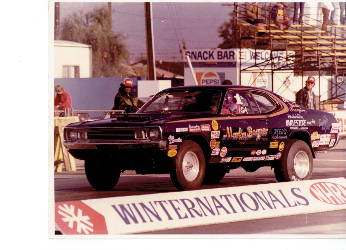 A black drag racing car with various sponsor decals performs a wheelie at a drag strip event. WINTERNATIONALS is visible on a barrier, with spectators and a snack bar in the background.
