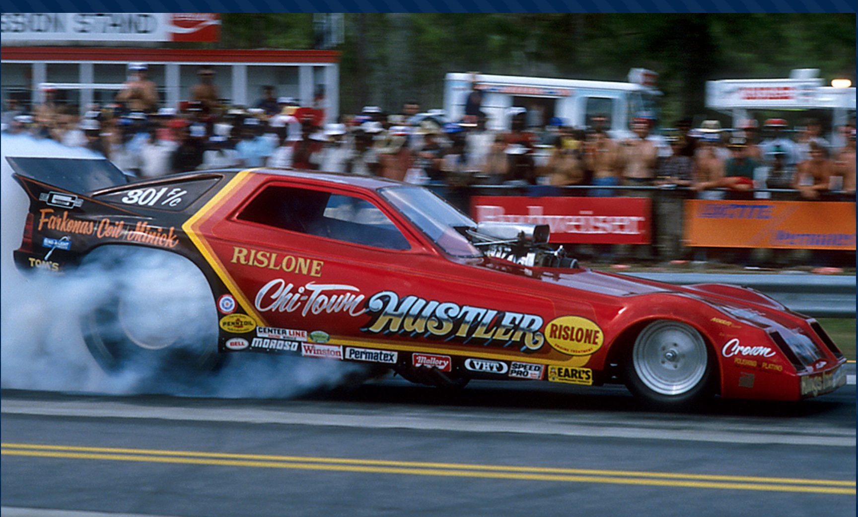 A red and yellow drag racing car with Chi-Town Hustler and sponsor logos does a burnout, creating smoke behind it, as spectators watch from behind a fence at a racetrack.