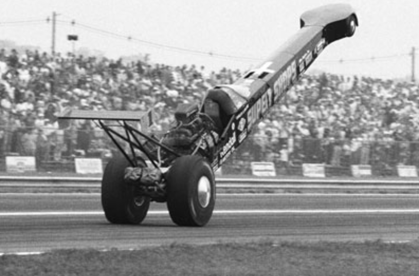 A drag racing car lifts its front end off the ground during a race on a track, with a large crowd of spectators watching in the background.