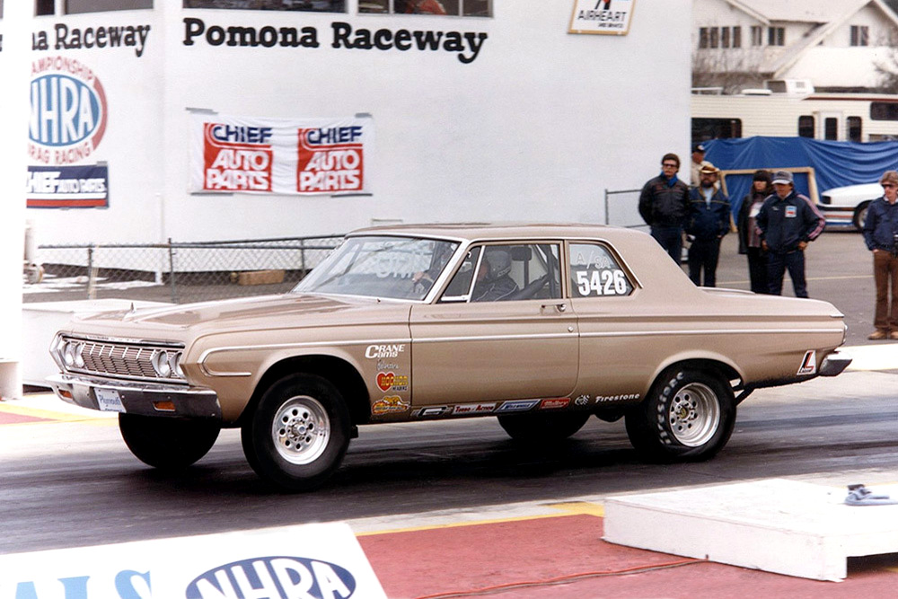 A vintage beige drag racing car with the number 5426 on the window is at the starting line of Pomona Raceway. Several people stand in the background near NHRA and auto parts signs.