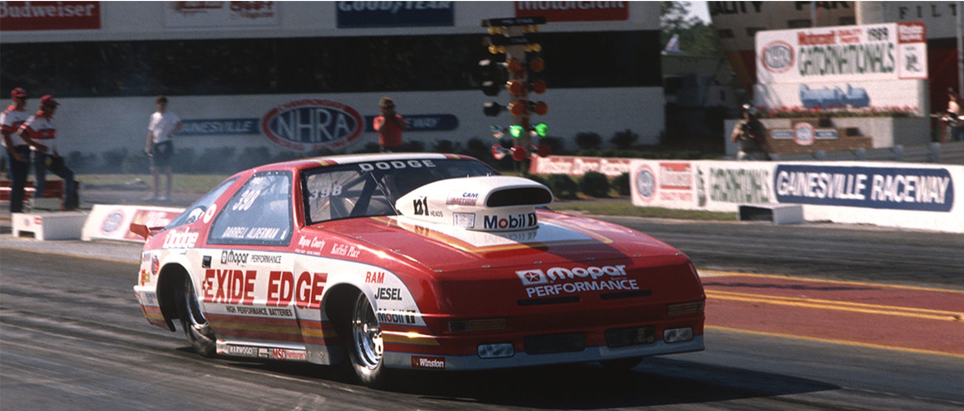 A red and white Dodge drag racing car speeds down the track at Gainesville Raceway, with sponsorship logos and crowds visible in the background. Racing lights and NHRA signage are also seen.