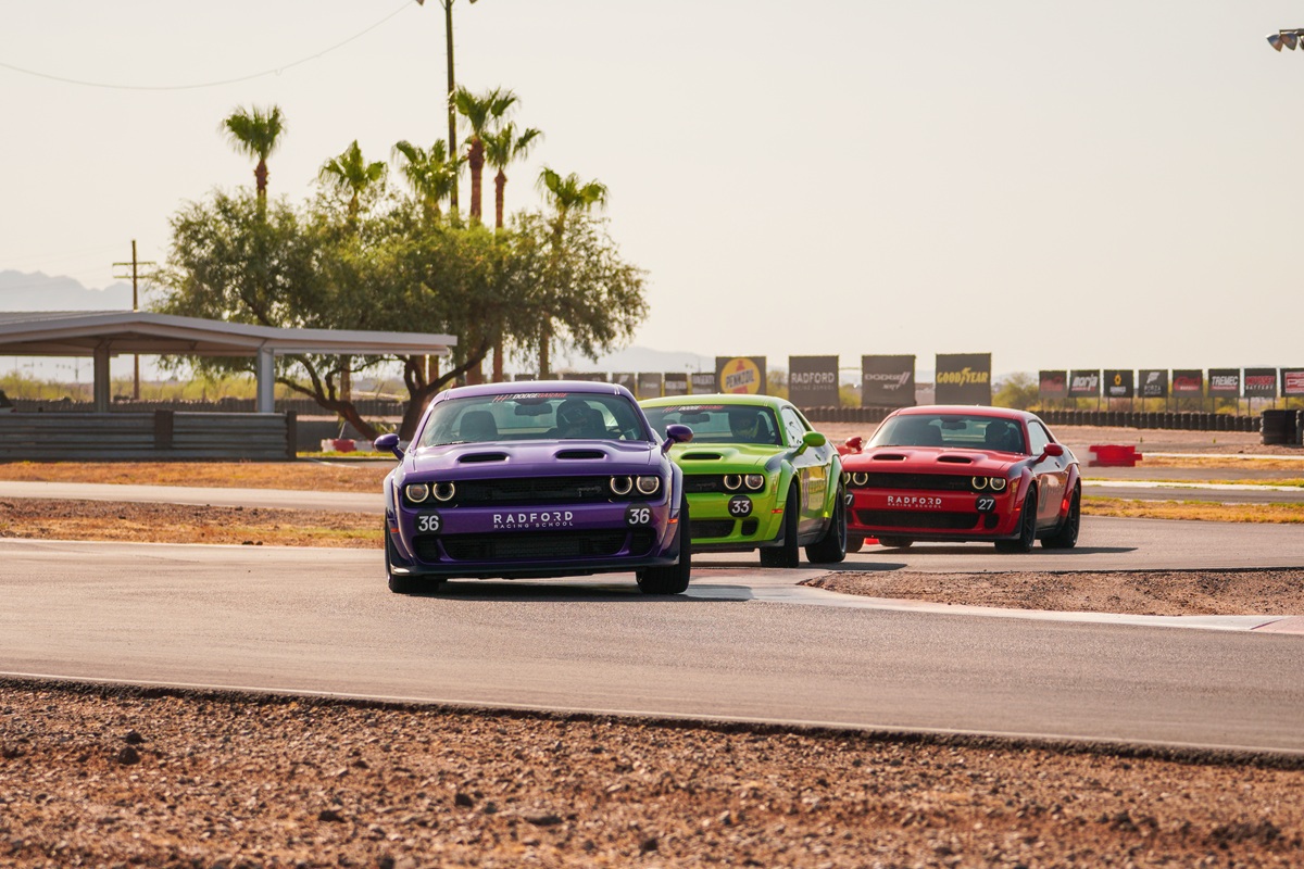 Three colorful Dodge sports cars—purple, green, and red—race closely on a sunny track, with palm trees and a covered area in the background. The cars are turning, and the scene suggests a lively, competitive atmosphere.