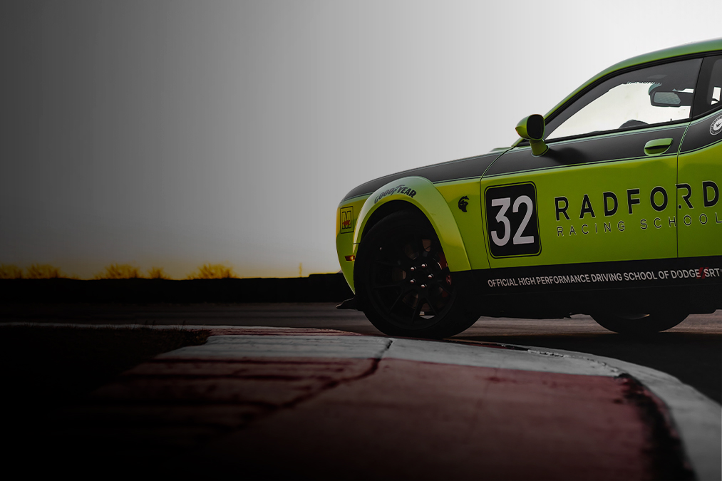 A green and black race car with number 32 and Radford Racing School branding is shown on a race track, partially visible from the side, with a blurred background and the sun low in the sky.