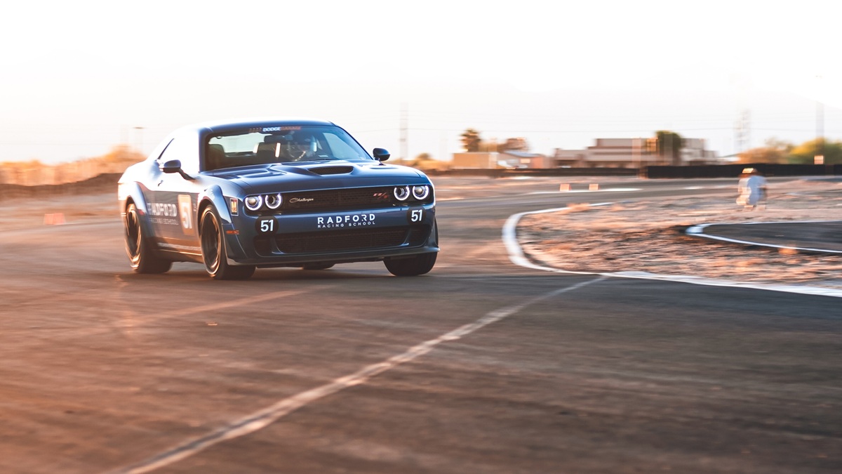 A black Dodge Challenger with Radford Racing School branding drifts on a racetrack, kicking up dust as it speeds through a curve under a clear sky.