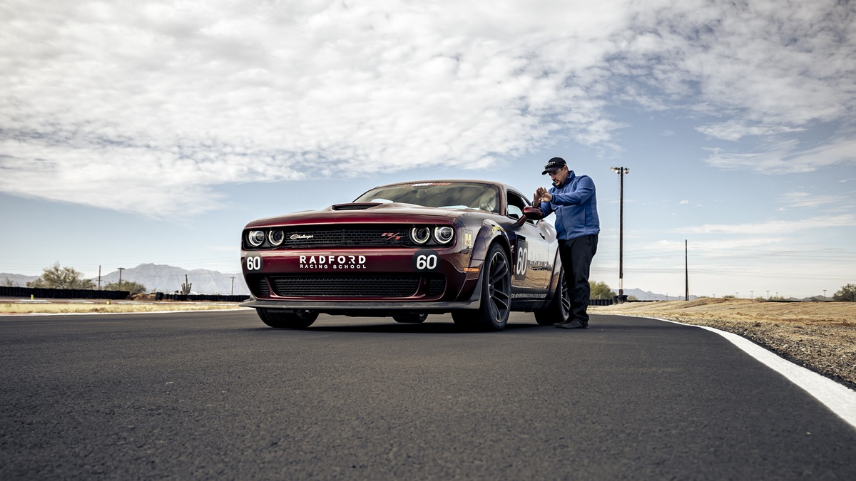 A person in a blue jacket and cap stands next to a dark red sports car on a racetrack under a partly cloudy sky, with mountains visible in the distance. The car has “Radford Racing School” on its front.