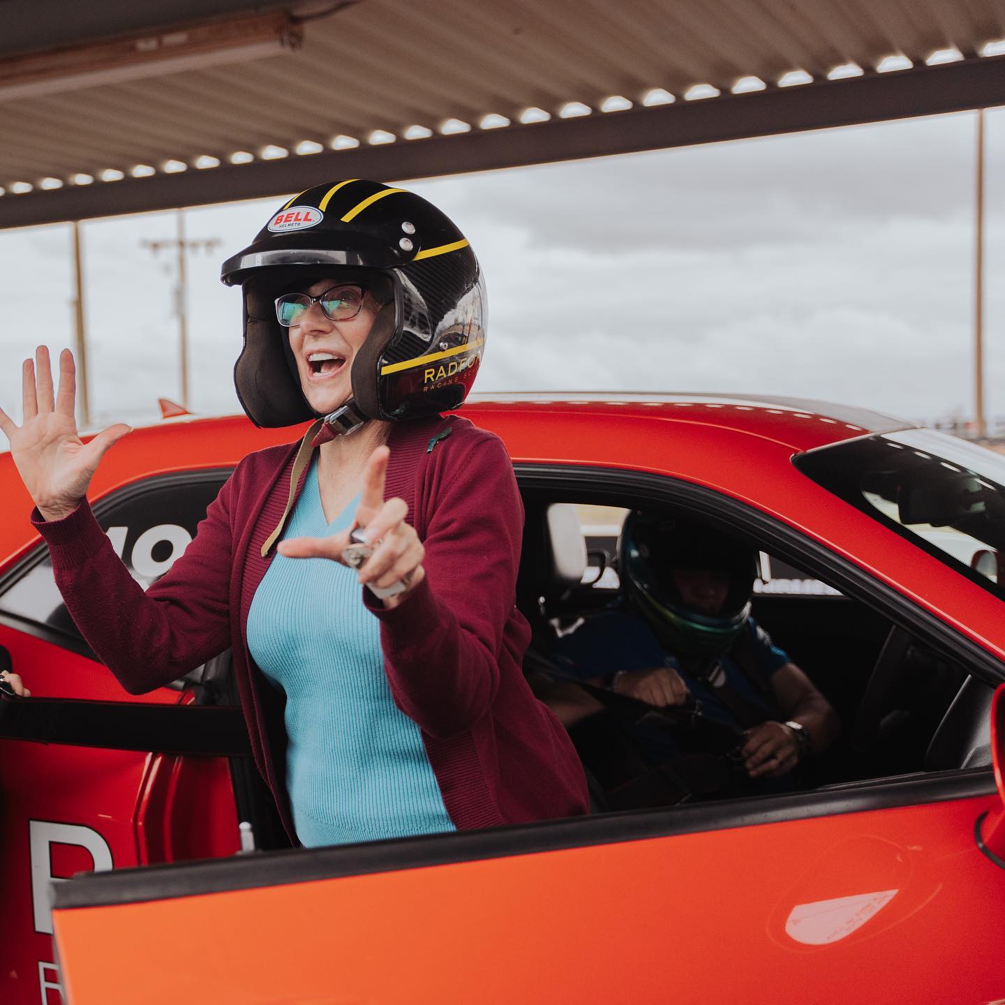 A woman wearing a racing helmet and glasses stands excitedly beside an orange sports car, holding up her hands and smiling. Another person sits inside the car, also wearing a helmet.