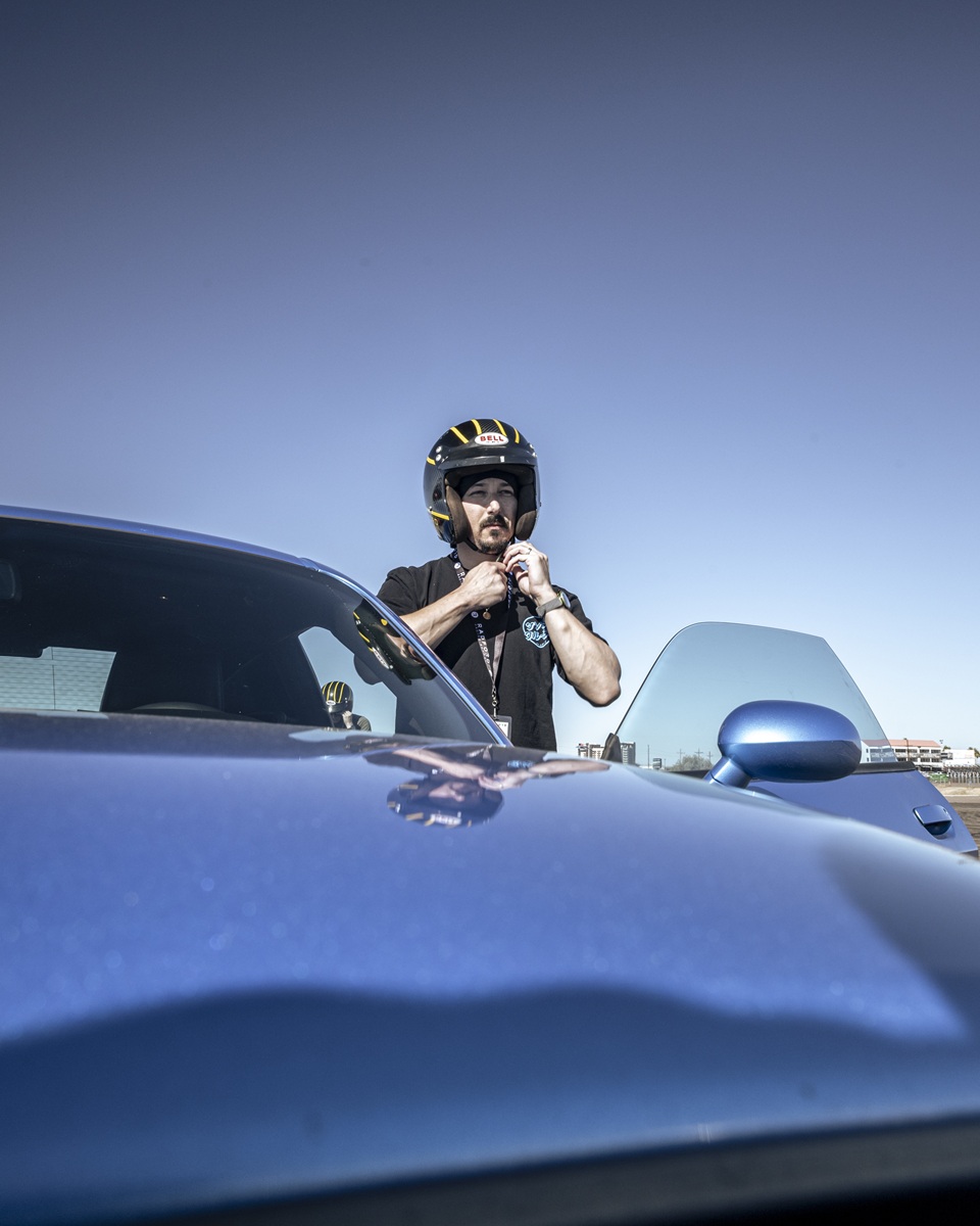A person wearing a helmet is standing next to a shiny blue sports car, fastening the helmet strap under their chin. The sky is clear and blue in the background.