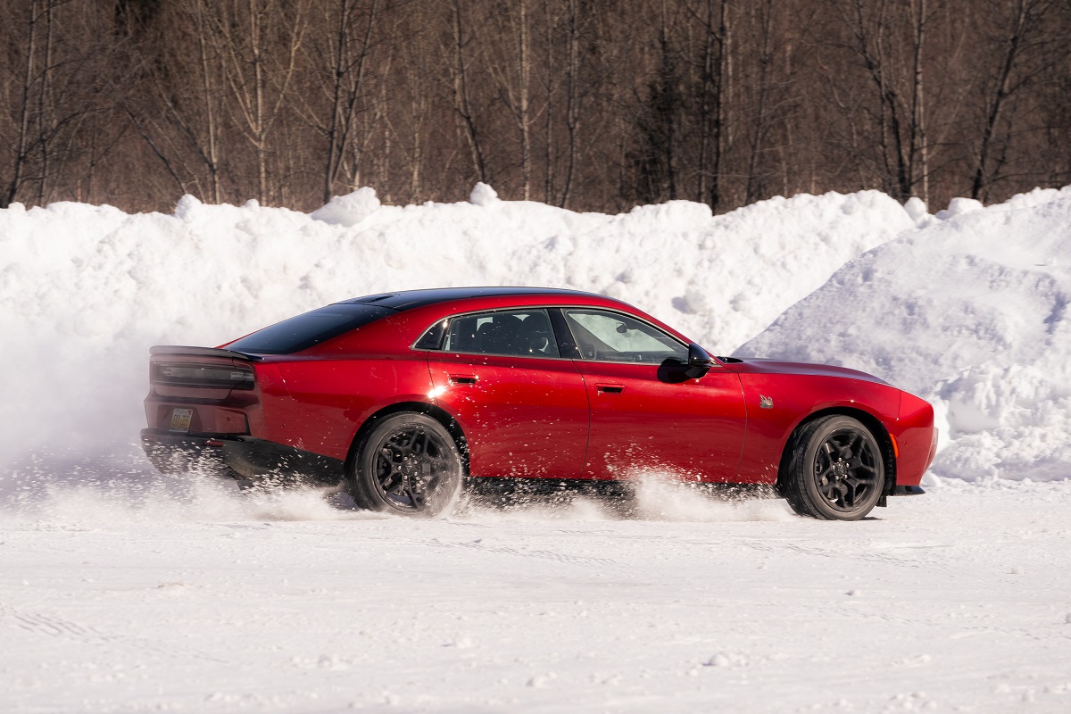A red sports car drives quickly across a snowy surface, kicking up snow behind its rear tires. Snowbanks and leafless trees are visible in the background.