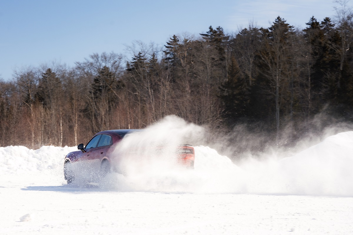 A red car driving fast on a snowy road kicks up a large spray of snow, with a forest of leafless trees in the background under a clear blue sky.