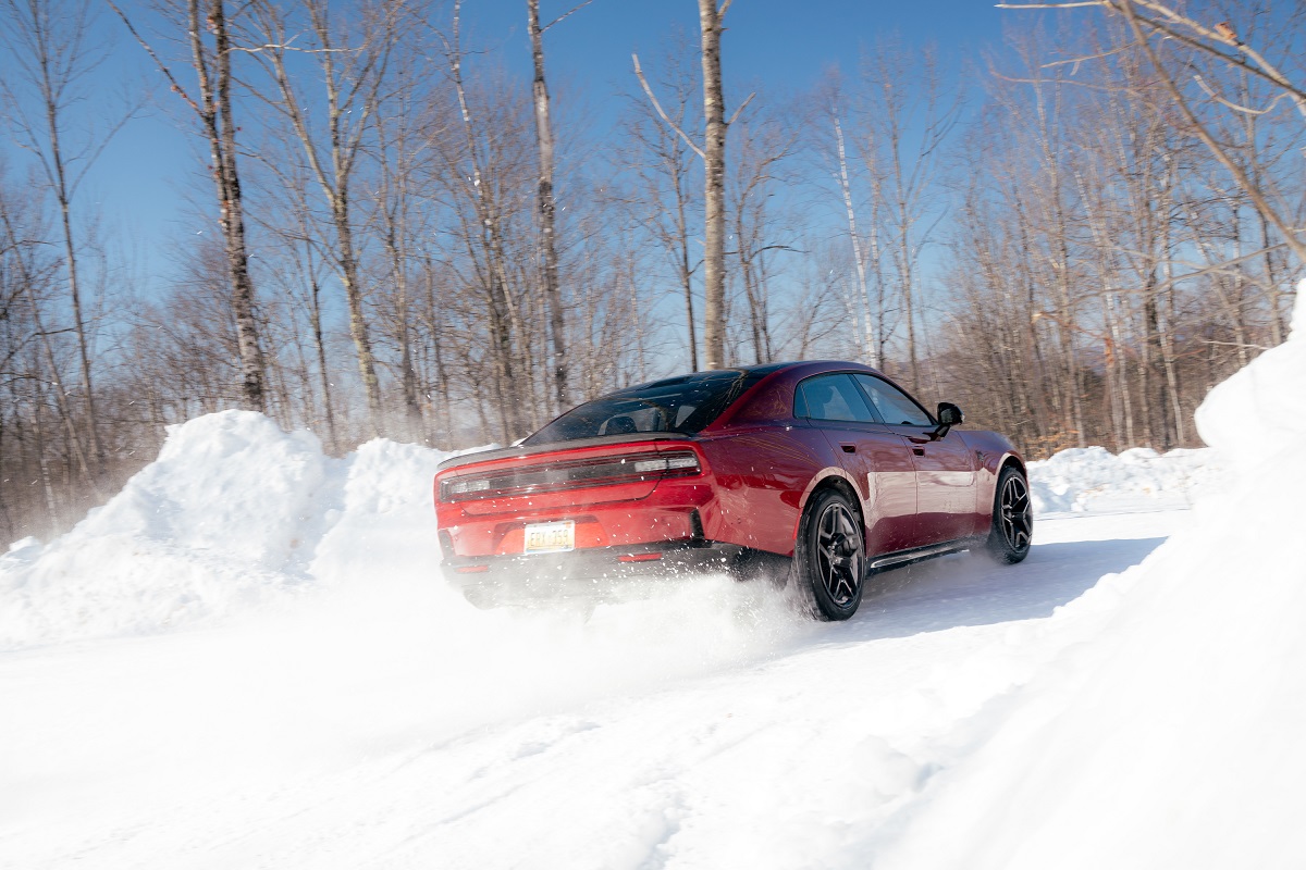A red sports car drives on a snow-covered road surrounded by snowbanks and leafless trees under a clear blue sky.