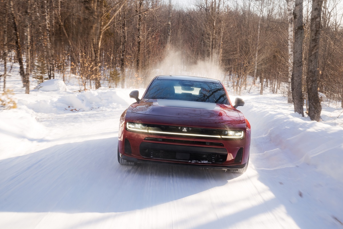 A red car drives quickly down a snow-covered road in a forested area, kicking up snow behind it. Bare trees line the road, and sunlight filters through, creating a crisp winter scene.