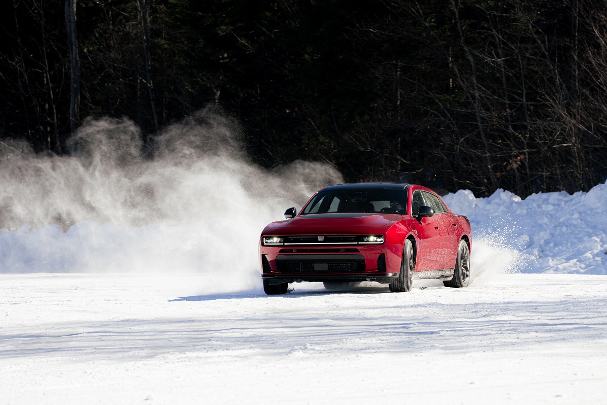 A red sports car drifts on a snow-covered road, kicking up a spray of snow, with trees and snowbanks in the background.