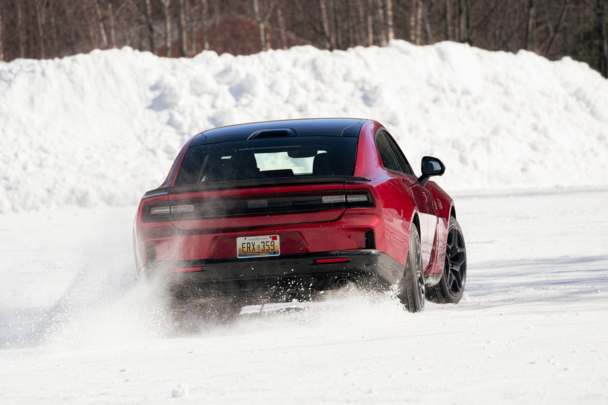 A red sports car drives on a snowy road, kicking up snow behind it. Tall snowbanks line the roadside, and leafless trees are visible in the background.