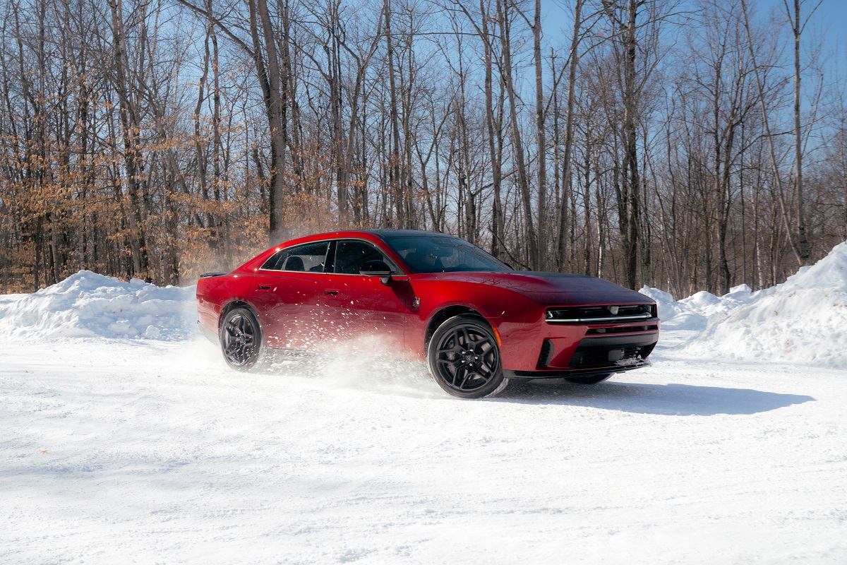A red sports car drives fast on a snowy road, kicking up snow, surrounded by snowbanks and bare trees in a winter landscape.