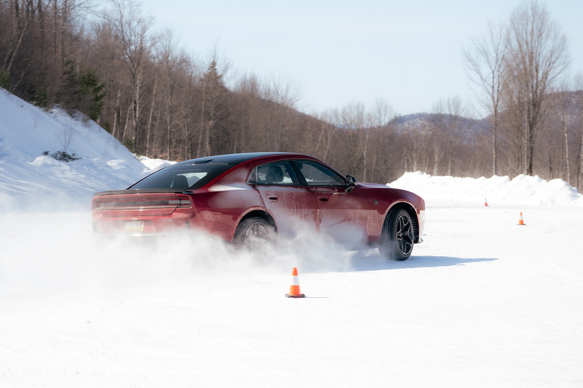 A red car skids and kicks up snow while maneuvering between orange cones on a snowy road, surrounded by leafless trees and snow-covered hills in the background.