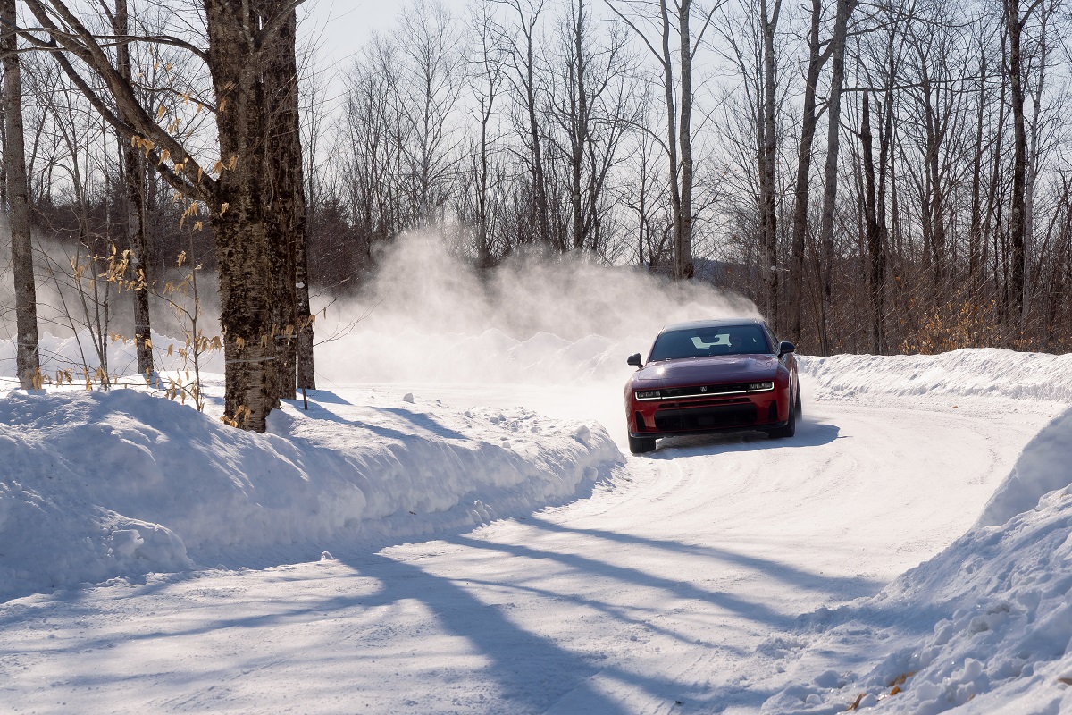A red car drives quickly around a snowy, curved road, kicking up snow behind it. Bare trees line the road, and sunlight casts shadows across the winter landscape.