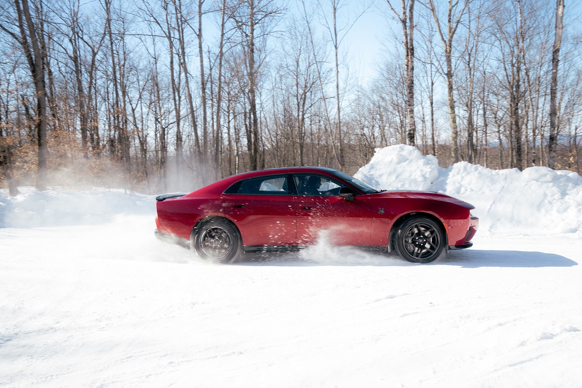 A red sports car drives quickly on a snowy road, kicking up snow with its tires. Bare trees and large snowbanks surround the road under a clear blue sky.