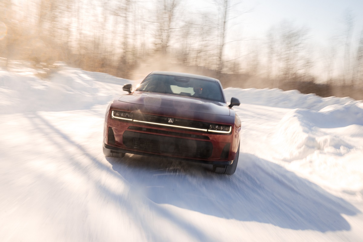A red car drives quickly through a snowy, winding road, kicking up snow with bare winter trees visible in the background under a bright sky.