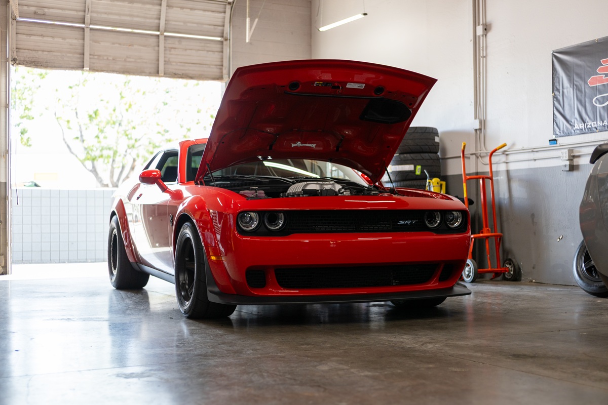 A red Dodge Challenger SRT with its hood open is parked inside a garage, with light coming in from an open door in the background. Various tools and tires are visible around the workspace.