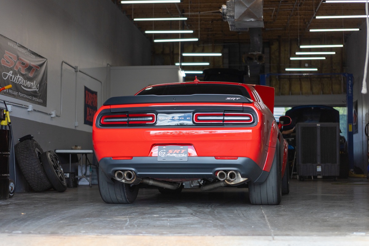 A red Dodge Challenger SRT parked inside a garage, viewed from the rear, with its taillights glowing and the hood open. The garage is well-lit and automotive equipment is visible on the sides.