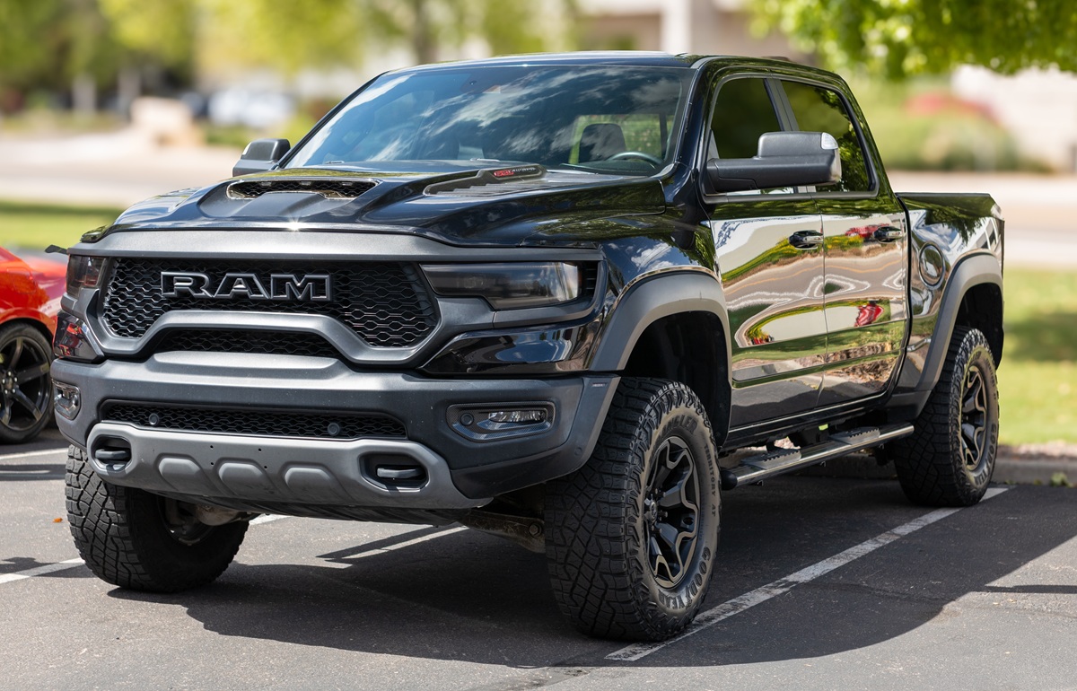 A black RAM pickup truck with rugged tires is parked in a lot on a sunny day, surrounded by trees and other vehicles.