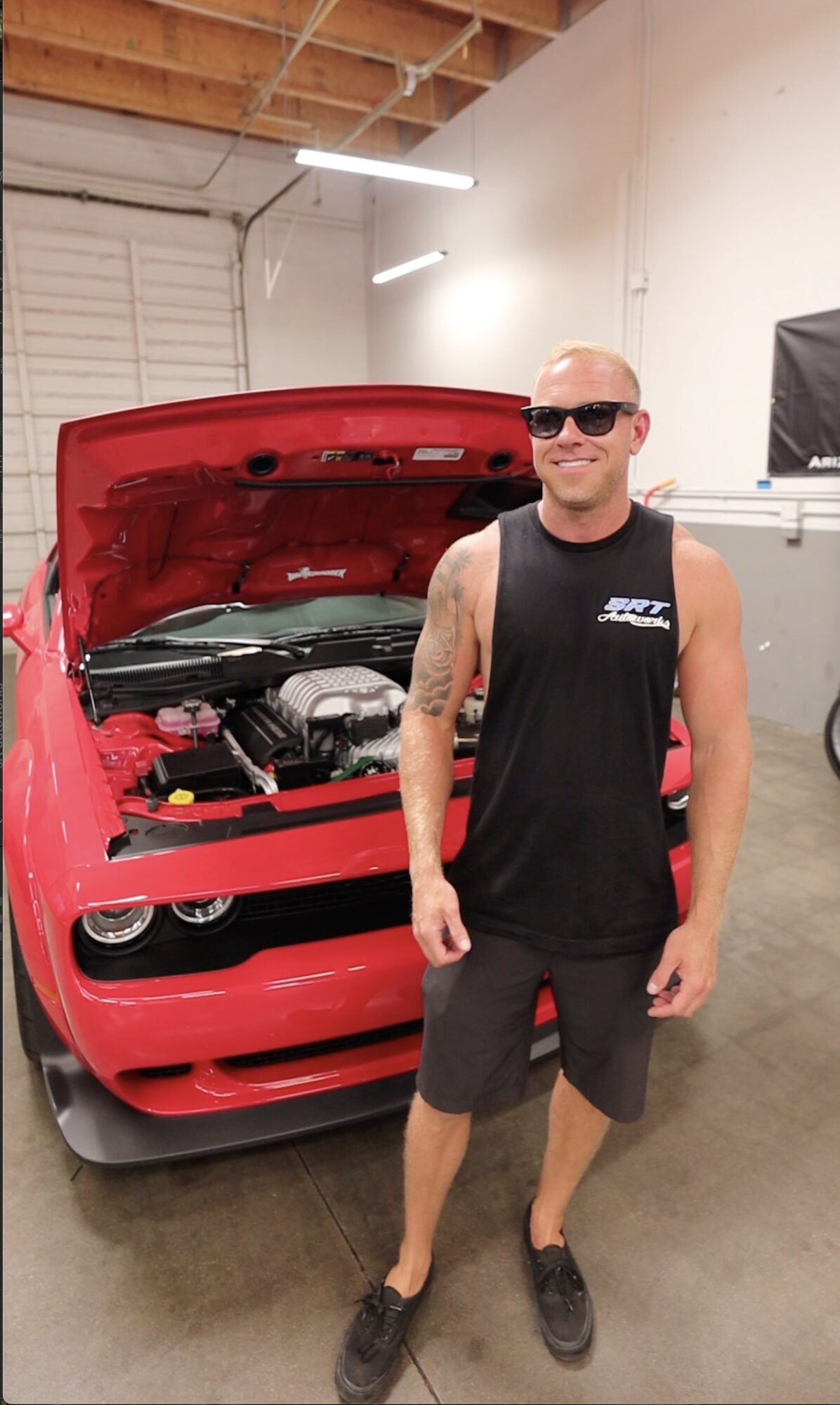 A man wearing sunglasses, a black tank top, and shorts stands smiling in front of a red sports car with its hood open inside a garage.