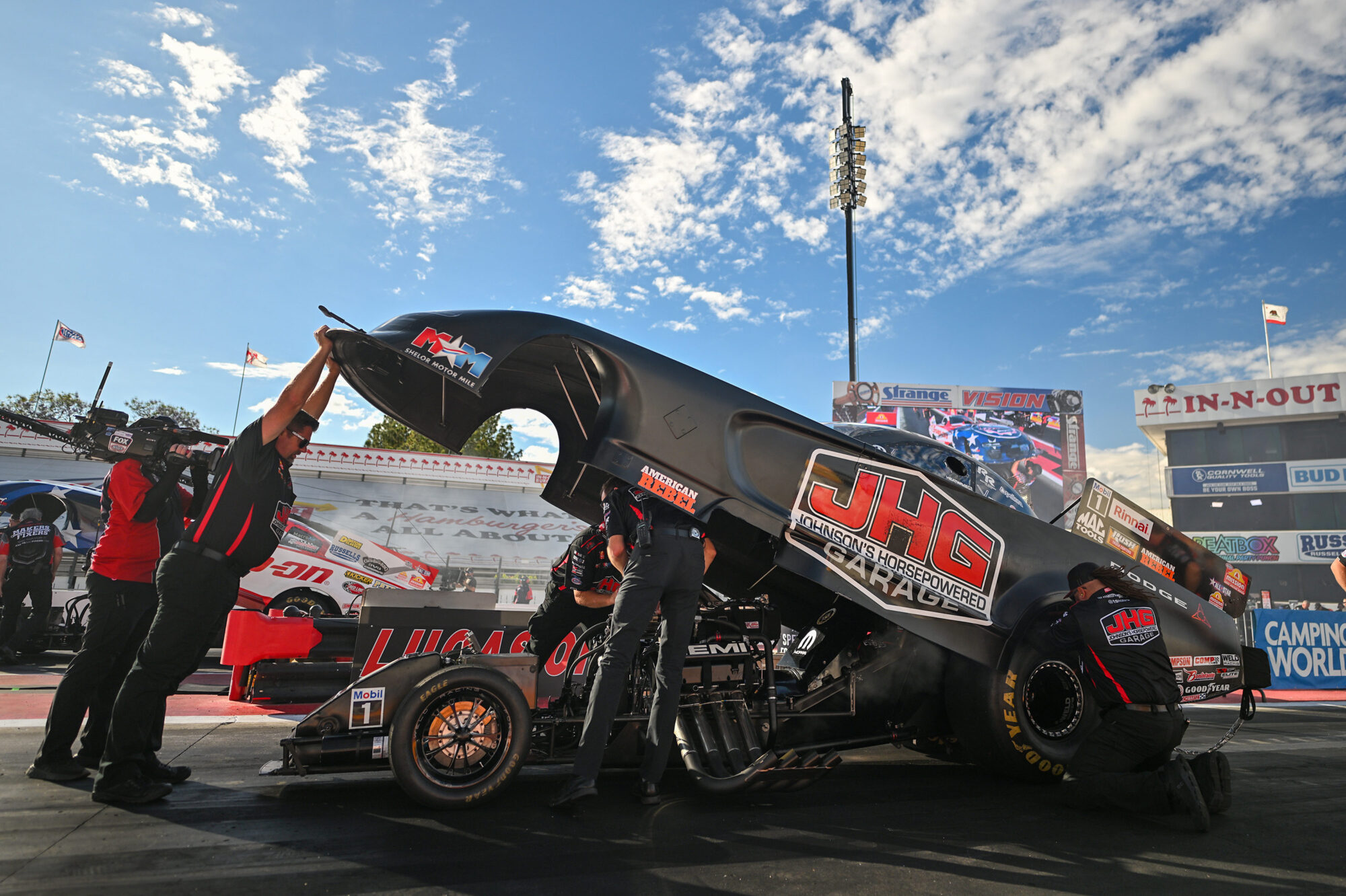 A team of mechanics works on a drag racing car with its body lifted, revealing the engine, at a racetrack under a partly cloudy sky. The car displays JHG and Lucas Oil logos, with grandstands and signage in the background.