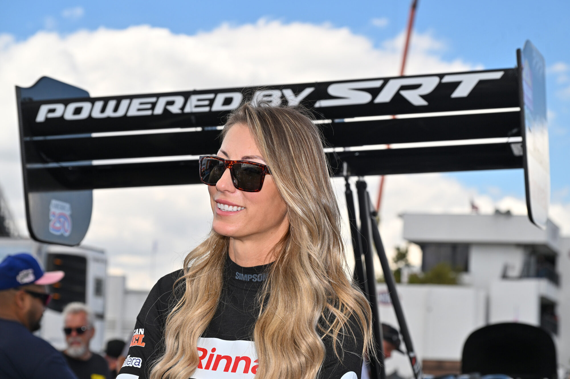 A woman with long blonde hair wearing sunglasses and a racing suit smiles in front of a large rear wing that reads “POWERED BY SRT” at a motorsport event. People and cars are visible in the background.