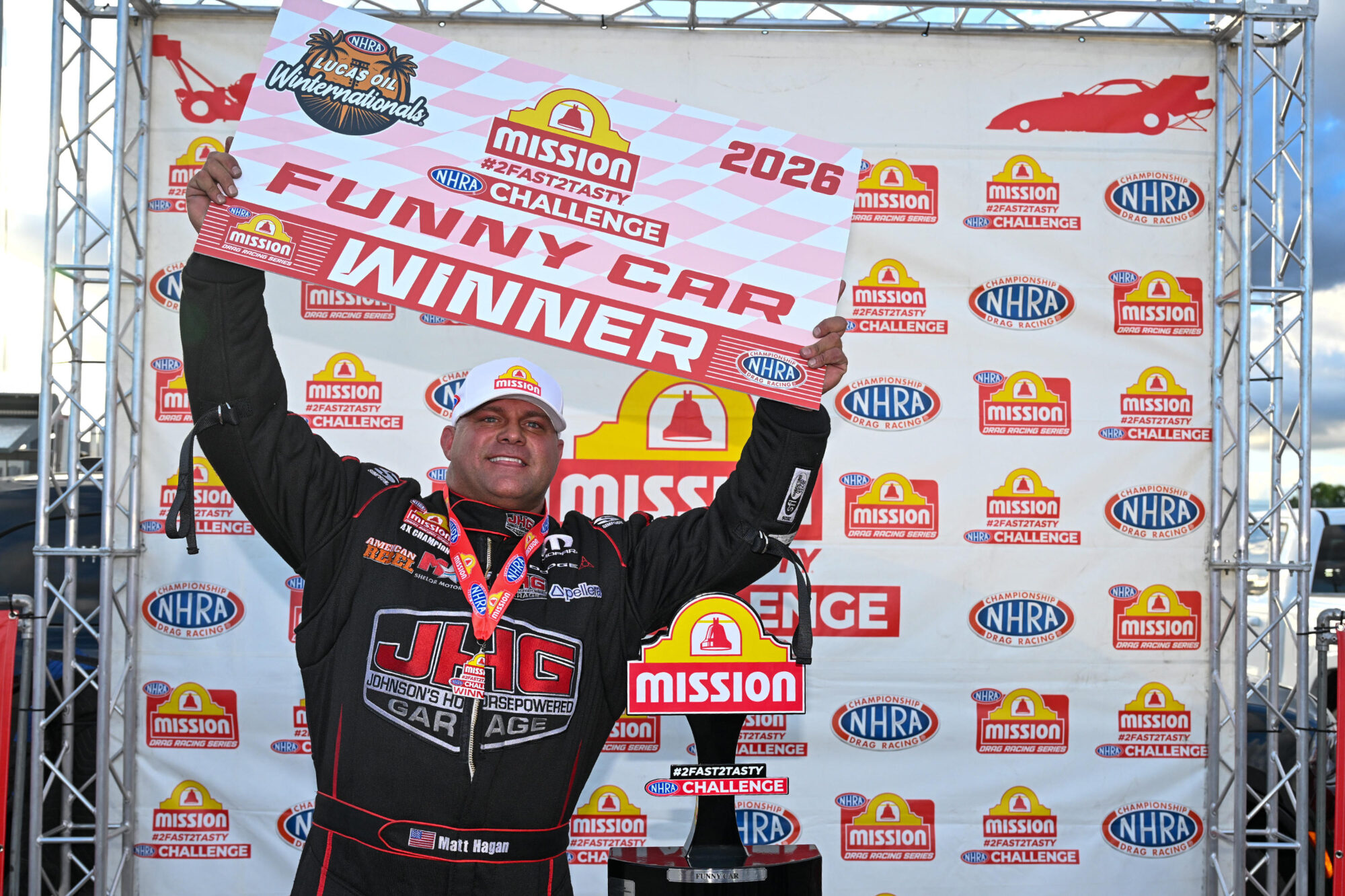 A race car driver in a black suit and cap holds up a Funny Car Winner sign and stands beside a trophy in front of a Mission Challenge winner’s backdrop at an NHRA event.
