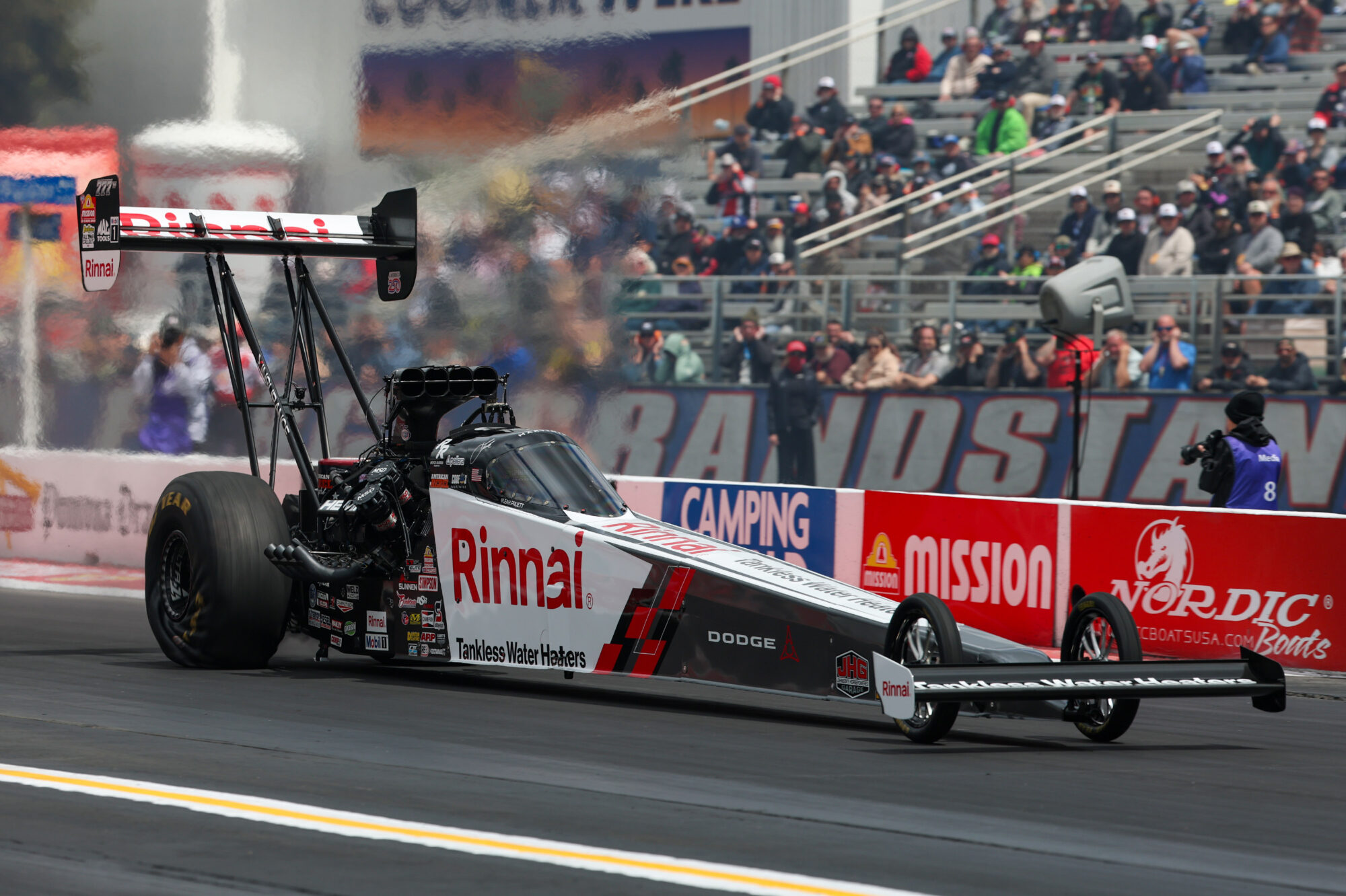 A dragster with a Rinnai sponsorship races down the drag strip, producing smoke from the rear tires. Spectators fill the grandstands in the background, and event banners line the track.