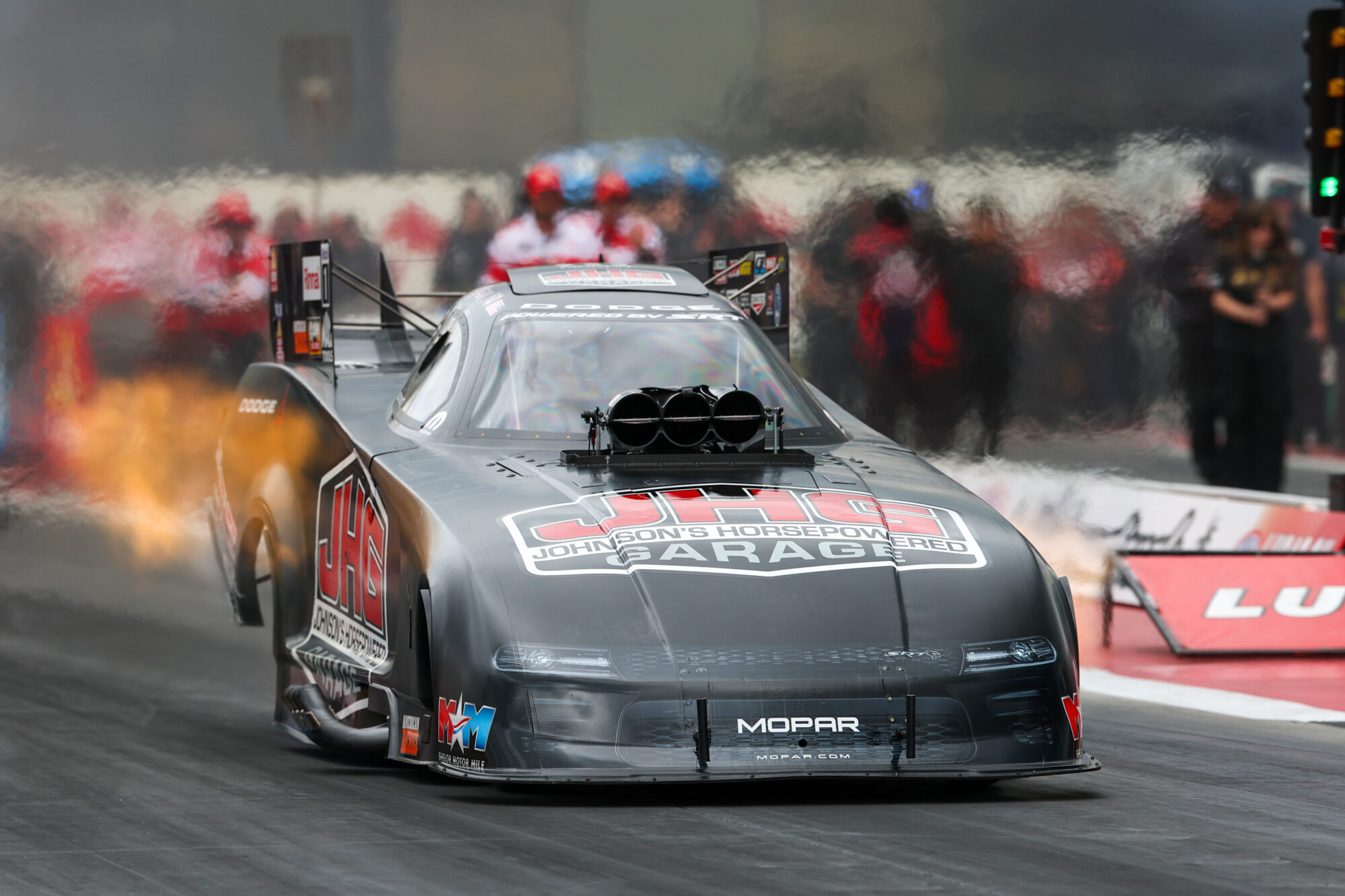 A black drag racing car with Johnsons Horsepowered Garage branding speeds down a dragstrip, flames visible from its exhaust, as crew members watch from the background.
