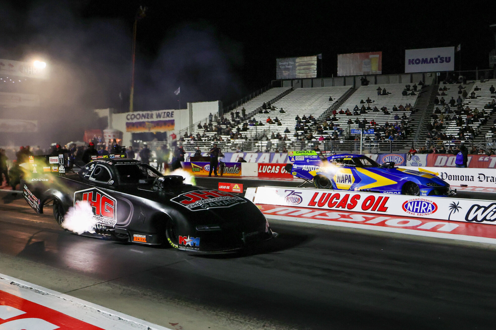 Two drag racing cars, one black with JHG branding and one blue with NAPA branding, accelerate side by side on a lit racetrack at night, with spectators in the grandstands in the background.