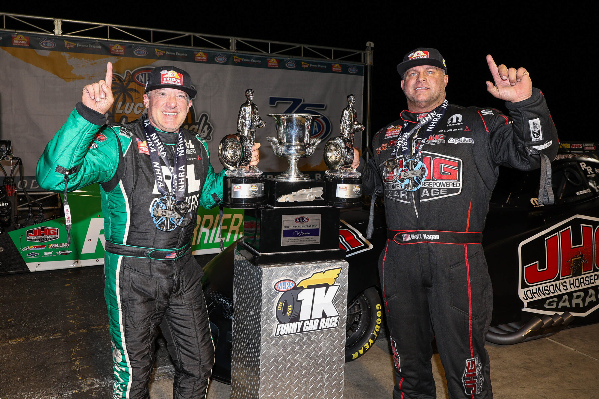 Two race car drivers in racing suits stand beside a large trophy and hold up one finger each, smiling and celebrating their victory at a nighttime motorsports event.