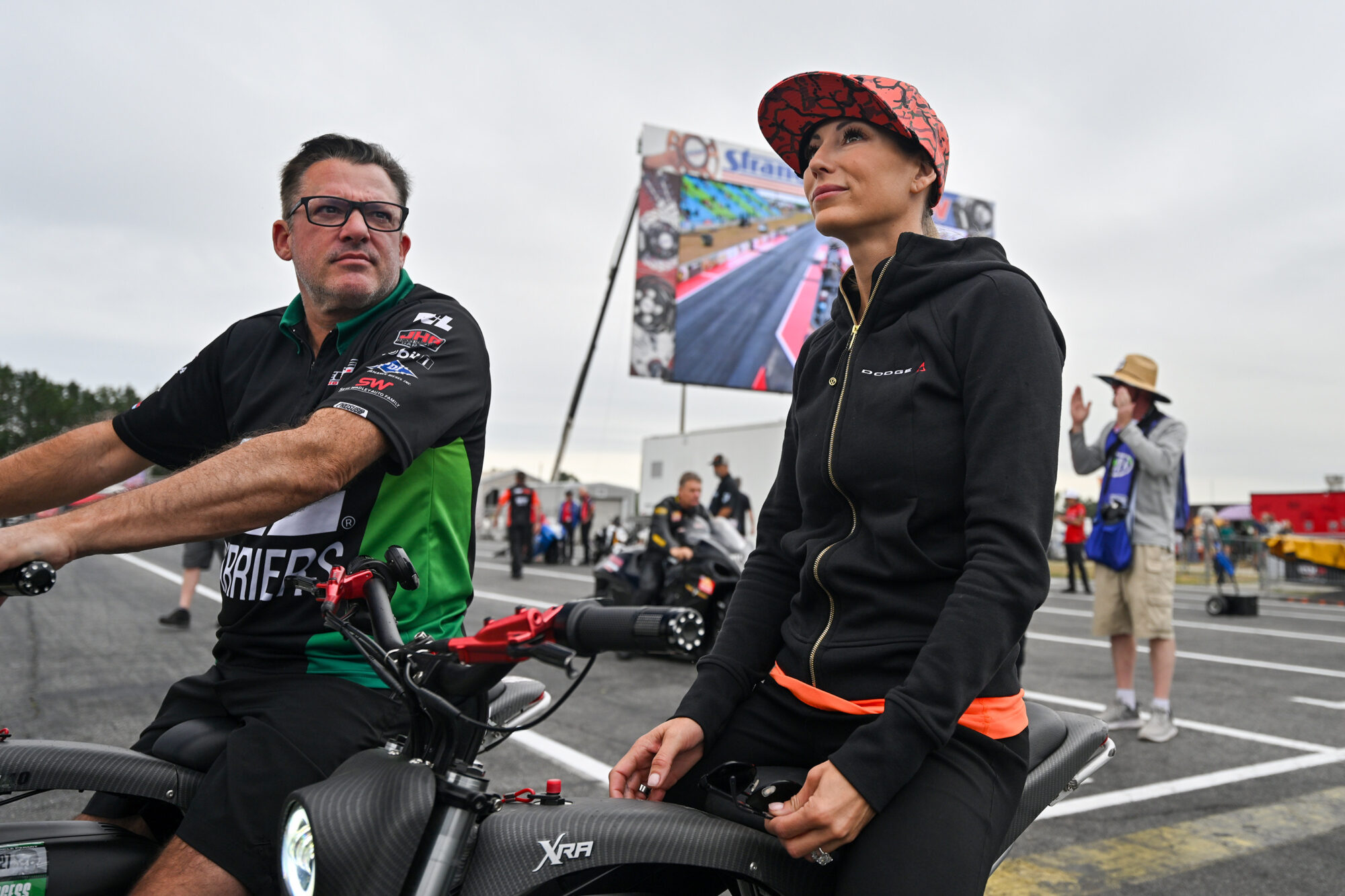 Two people sit on electric bikes at a race track. The woman wears a patterned cap and black jacket, while the man wears glasses and a black and green shirt. A large screen and several people are visible in the background.
