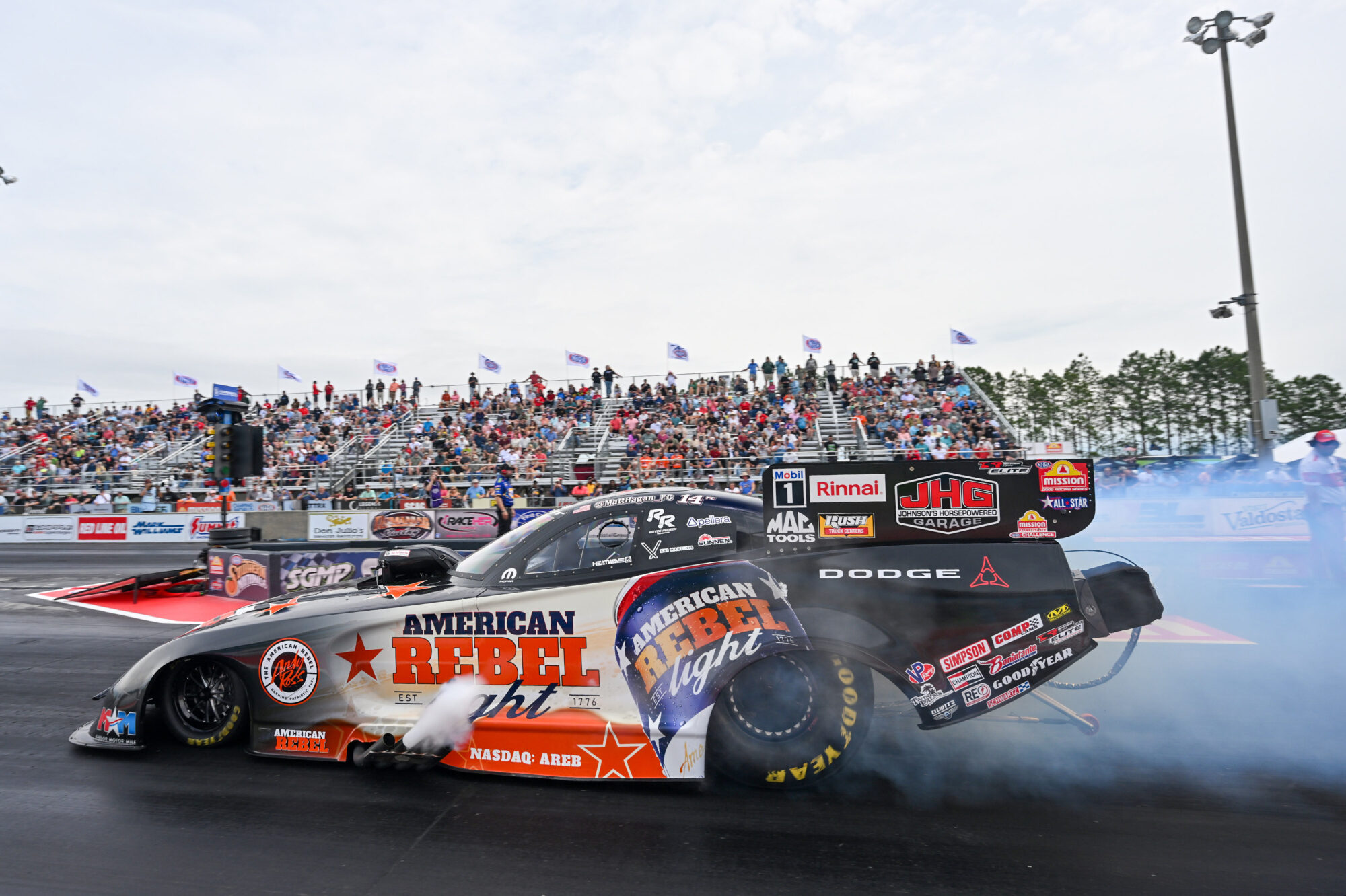 A drag racing car with American Rebel branding burns rubber at the start line, producing smoke. Spectators fill the grandstands in the background, watching the event under a partly cloudy sky.