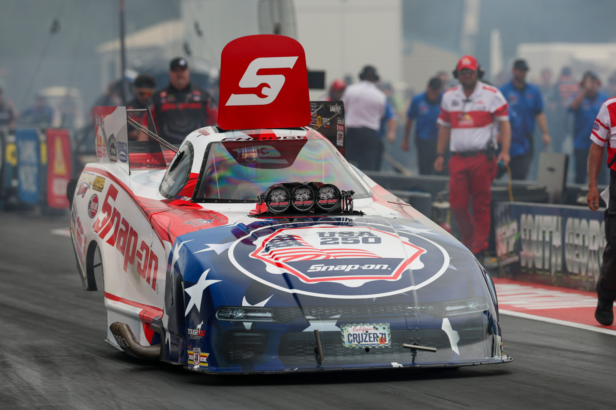 A patriotic-themed drag racing car with Snap-on branding lines up on the track, surrounded by crew members in racing uniforms, ready for a race.