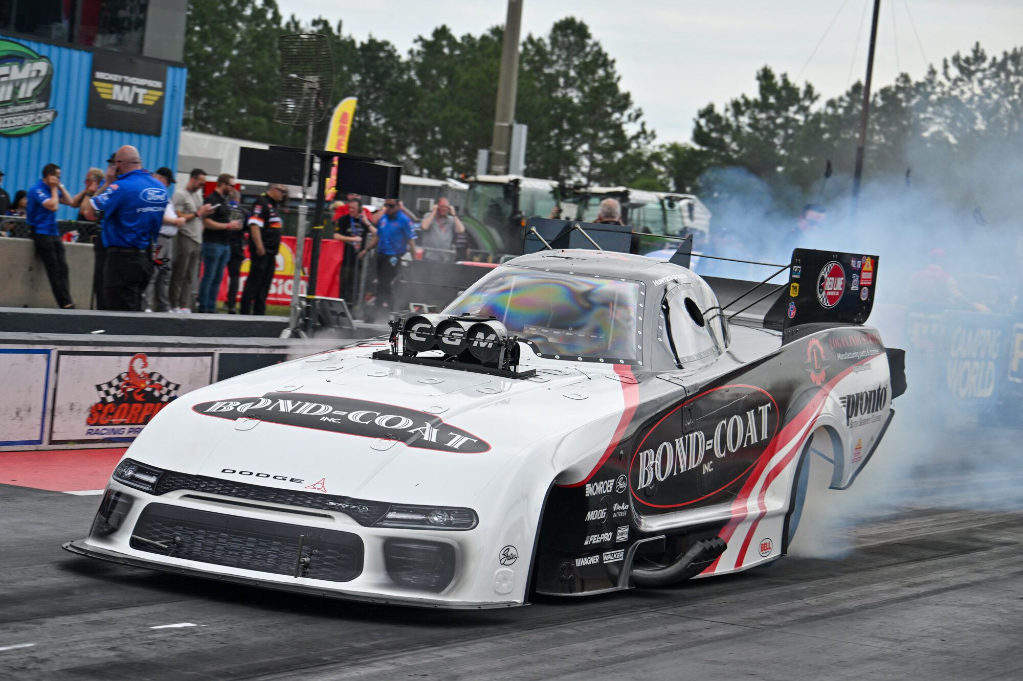 A black and white Dodge drag racing car with Bond-Coat branding emits smoke from its tires as it accelerates on a racetrack, with crew members and spectators in the background.