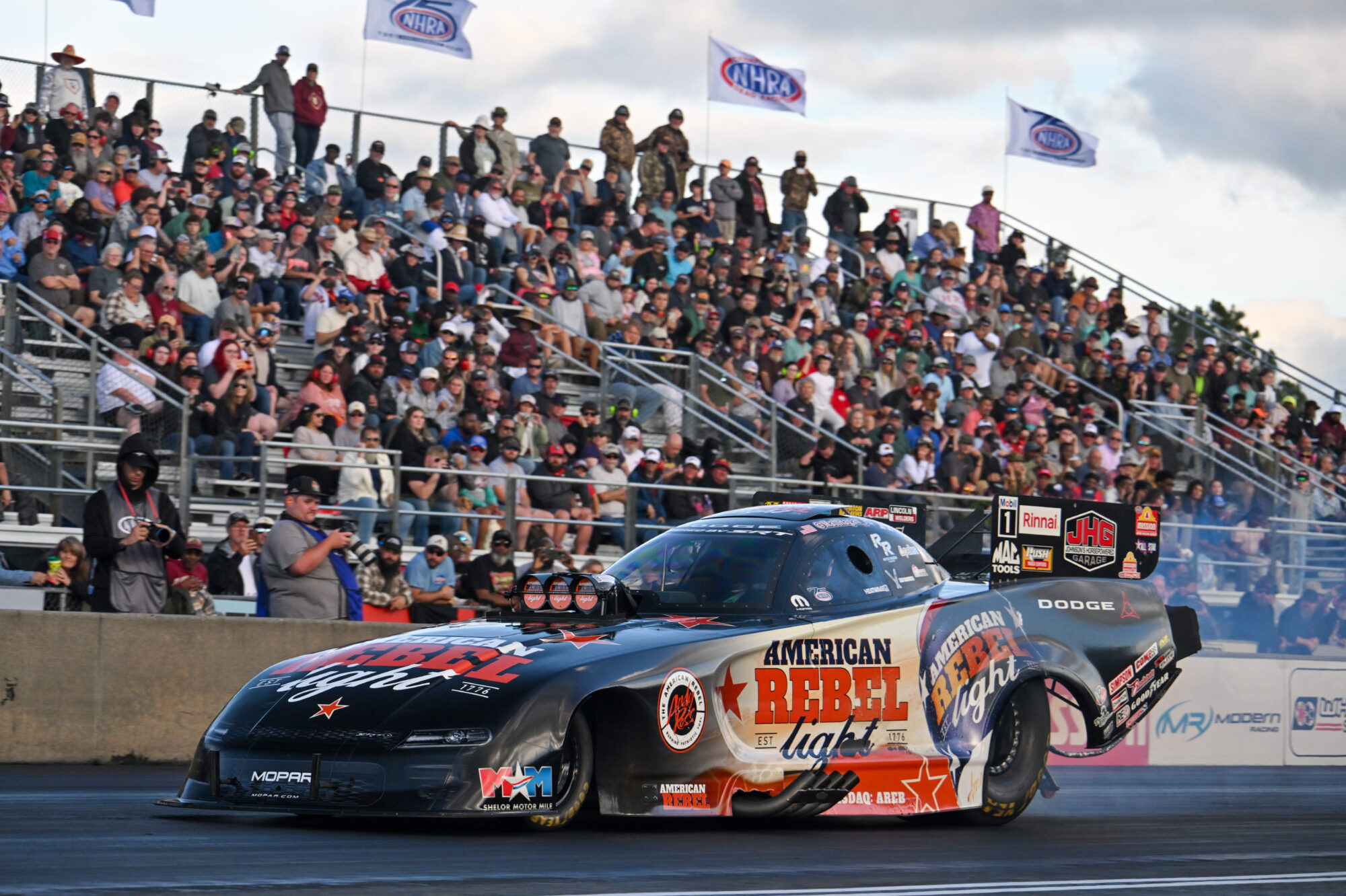 A black drag racing car with American Rebel branding speeds down a track as a large crowd watches from bleachers; NHRA flags are visible in the background.