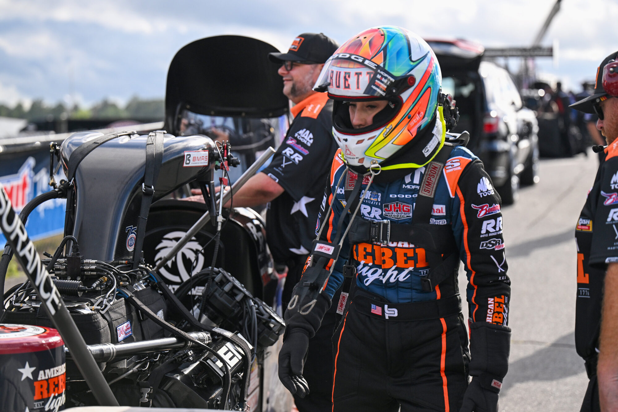 A race car driver in a colorful helmet and full racing suit stands beside a dragster, with team members nearby. The scene is set on a racetrack with cars and equipment in the background.