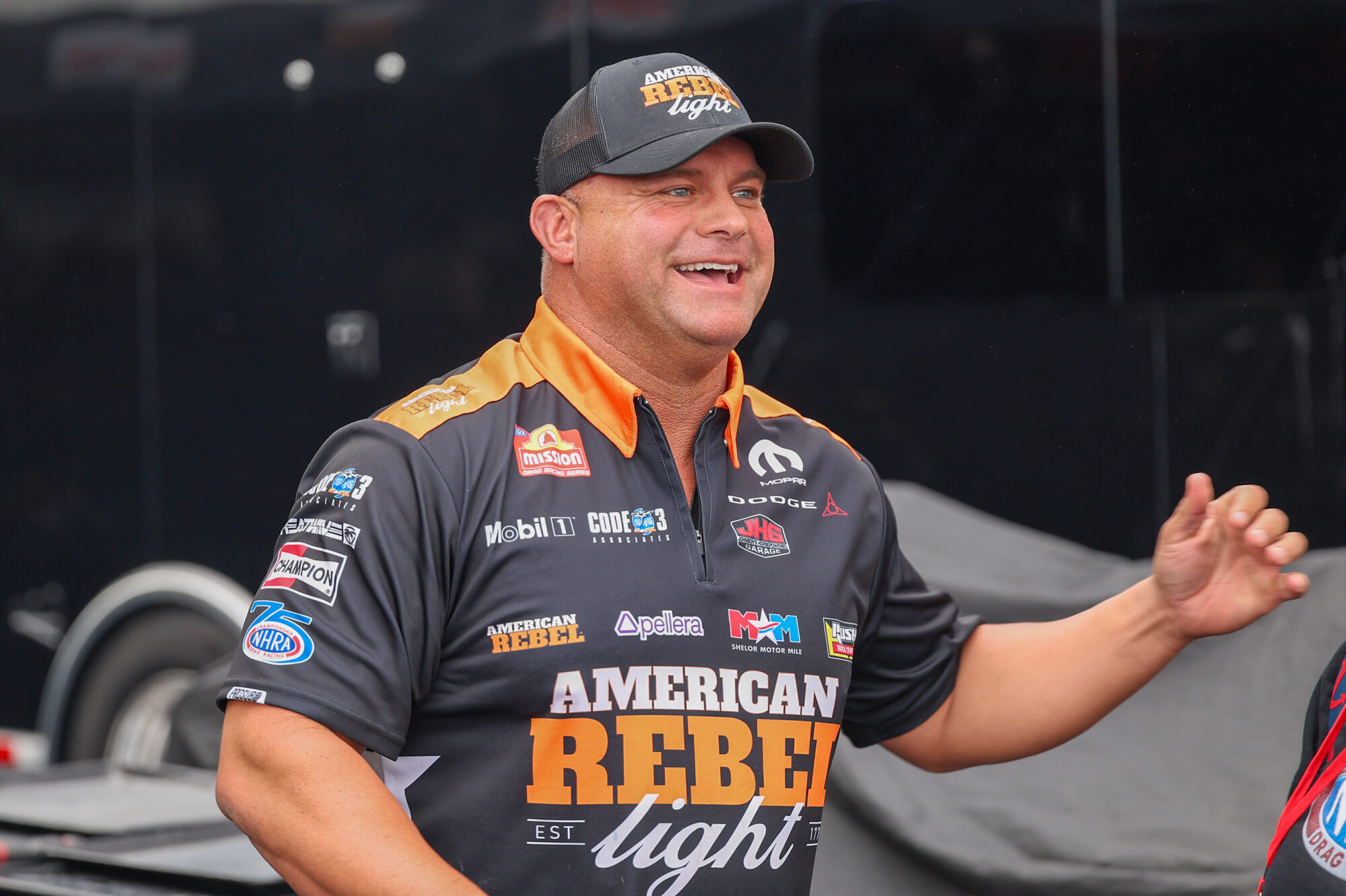 A smiling man wearing a black American Rebel Light racing shirt with various sponsor logos stands with his arm slightly raised in front of a dark background.