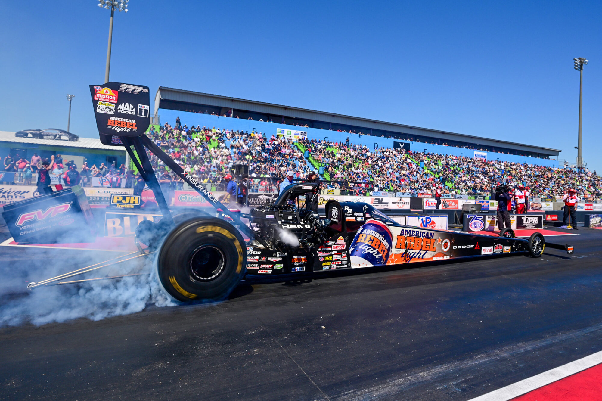 A dragster car burns rubber at the starting line of a racetrack, creating smoke, with a crowd of spectators watching from blue and green bleachers under a clear blue sky.