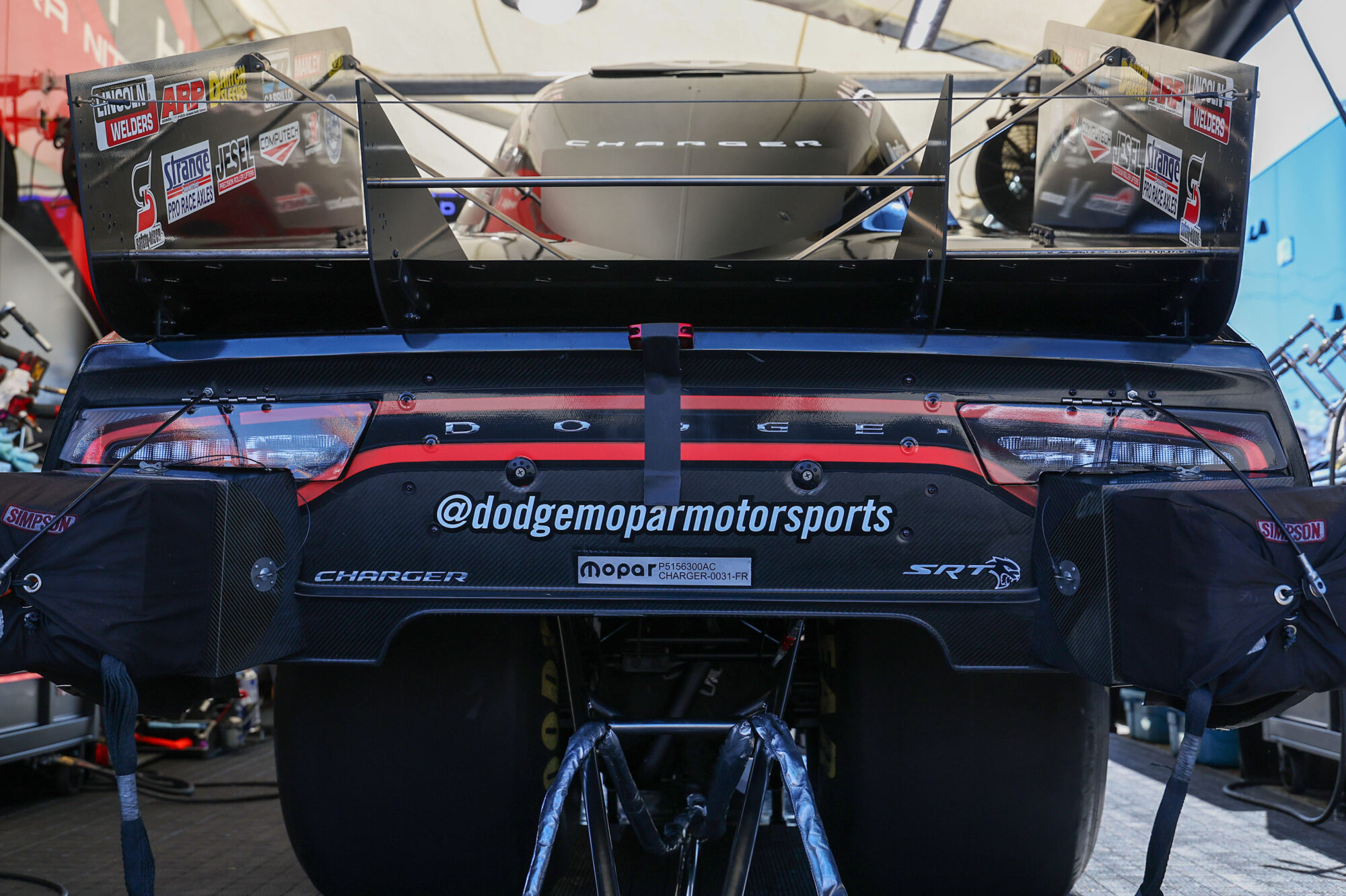 Close-up view of the rear end of a Dodge Charger racing car in a pit area, showing aerodynamic components, sponsor logos, and the Instagram handle @dodgemoparmotorsports prominently displayed on the bumper.