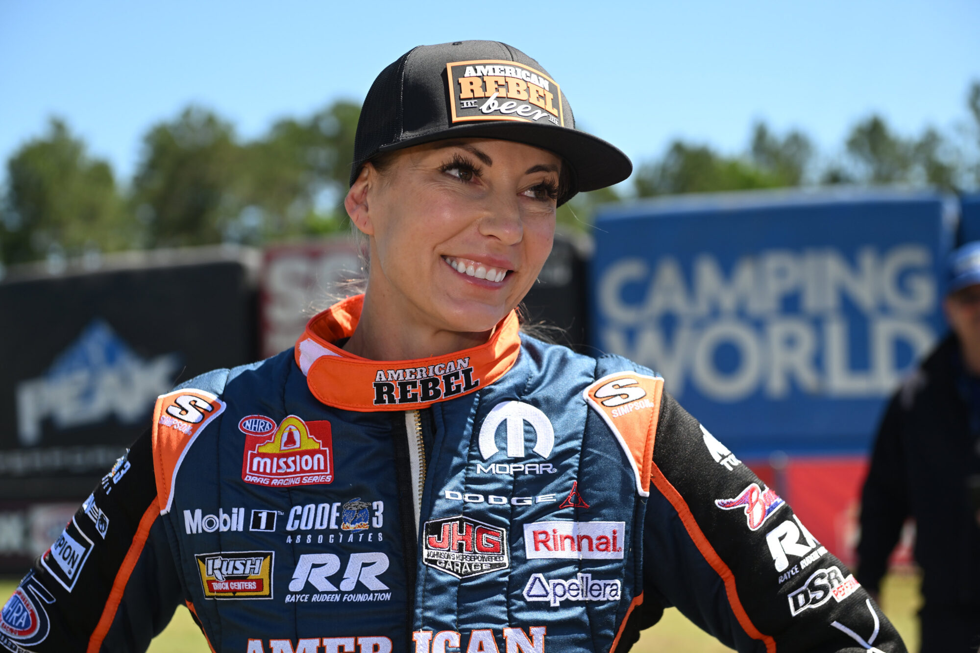 A smiling female race car driver in a blue and orange racing suit and a black “American Rebel” cap stands outdoors with a racing track and sponsor banners in the background.